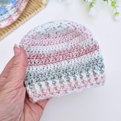 A hand holding a small crocheted beanie hat with horizontal stripes in white, dusty pink, and light gray, set against a bright white background with soft decorative elements.