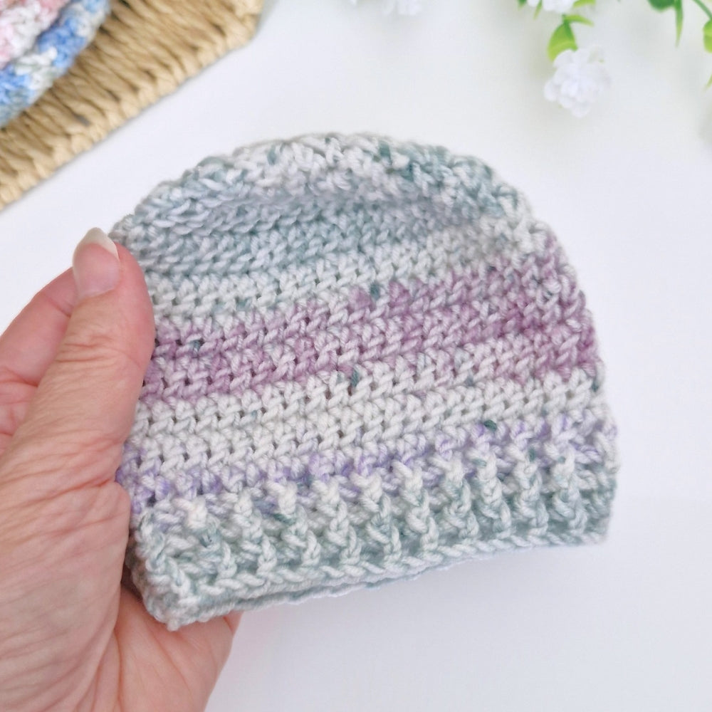 A hand holding a small handmade crocheted beanie hat with horizontal stripes of light gray, sage green, and lavender purple, set against a bright white background with soft decorative elements.