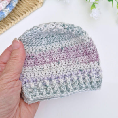 A hand holding a small handmade crocheted beanie hat with horizontal stripes of light gray, sage green, and lavender purple, set against a bright white background with soft decorative elements.