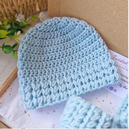 Blue crochet baby hat and booties on a white surface with a cardboard box in the background.