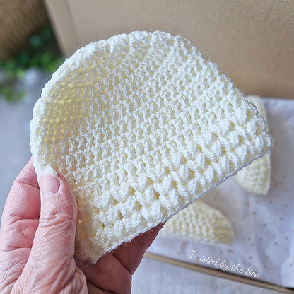 Cream crocheted baby hat held by a hand with a blurred background