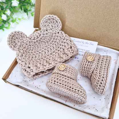 A three-piece crochet baby bootie set in a kraft gift box. The set includes a soft knit hat with bear ears, a pair of soft knit booties, in praline paradise. 