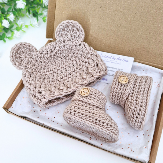 A three-piece crochet baby bootie set in a kraft gift box. The set includes a soft knit hat with bear ears, a pair of soft knit booties, in praline paradise. 