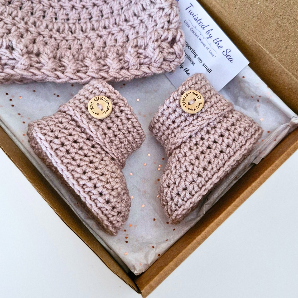 A handmade baby bootie set in a gift box, including a pair of dusty pink crocheted booties with gold buttons, a matching hat, and tissue paper. The brand name 'Twisted by the Sea' is visible on the tag.