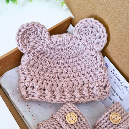 A handmade crochet baby hat with bear ears in a dusty pink color, displayed inside a brown cardboard gift box with a matching pair of booties and a tissue paper.