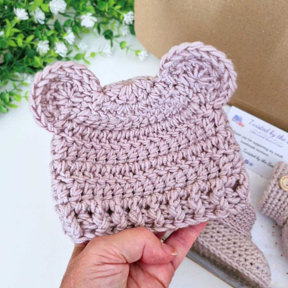 A hand holds a dusty pink crocheted baby hat with bear ears, shown against a styled background with a brown kraft box, white flowers, and matching booties in the background