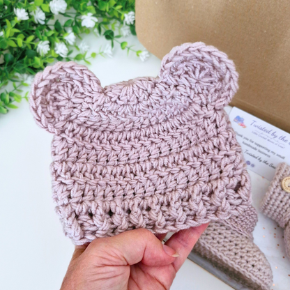 A hand holds a dusty pink crocheted baby hat with bear ears, shown against a styled background with a brown kraft box, white flowers, and matching booties in the background
