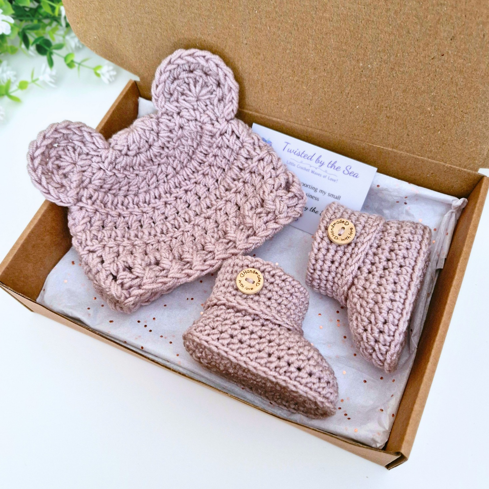 A handmade crochet baby gift set in a brown cardboard box, featuring a dusty pink knit hat with bear ears, matching booties, with a small card reading 'Twisted by the Sea'.