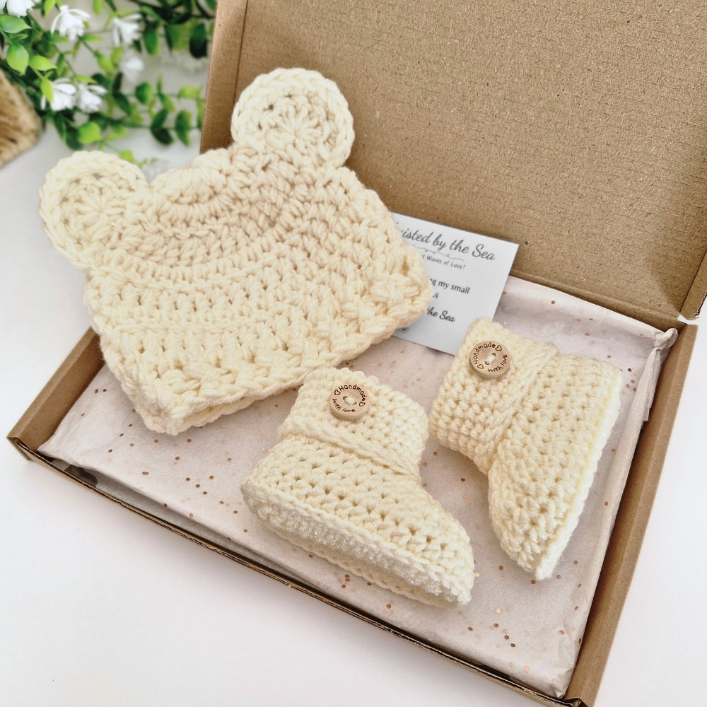 A handmade crochet baby gift set in a kraft cardboard box, featuring a cream-colored crochet hat with bear ears, a matching bootie, and a small card that reads 'Twisted by the Sea'.