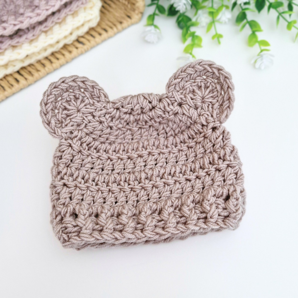 A handmade crochet baby hat in a dusty taupe-brown color, featuring a textured knit pattern and two small bear ears on top. It is displayed on a clean white surface with a woven mat and green foliage in the background.