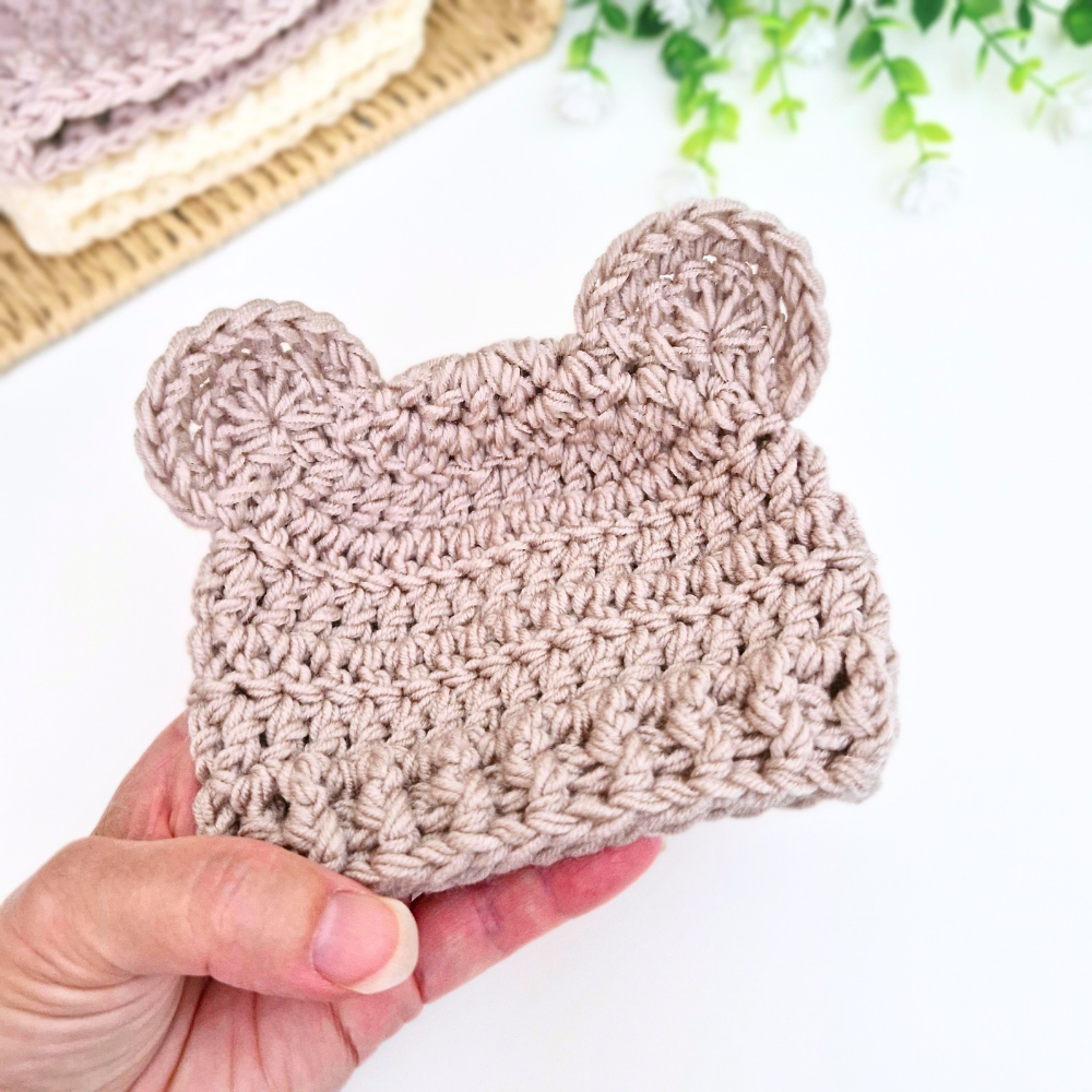 A hand holds a small crocheted beanie with bear ears in a light taupe color, with a second matching beanie stacked on a woven mat in the background.