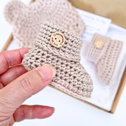 A hand holds a small beige crocheted baby bootie with a wooden button that reads 'Handmade with Love' in front of a box containing matching booties.