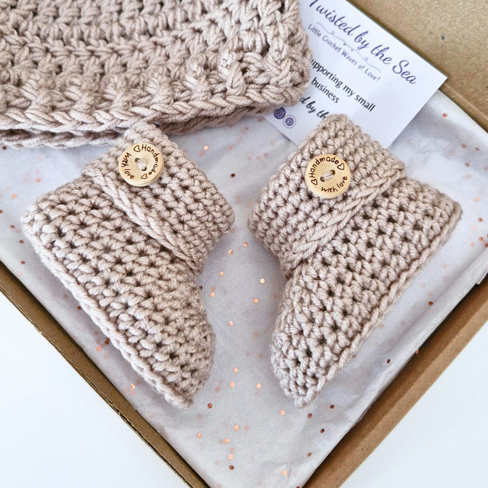 A handmade baby bootie set in a kraft gift box, featuring a pair of crochet beige booties with wooden buttons and a matching knit hat. The packaging includes a branded card from 'Twisted by the Sea'.