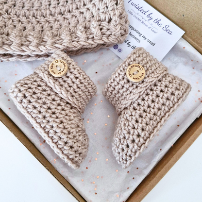 A handmade baby bootie set in a kraft gift box, featuring a pair of crochet beige booties with wooden buttons and a matching knit hat. The packaging includes a branded card from 'Twisted by the Sea'.