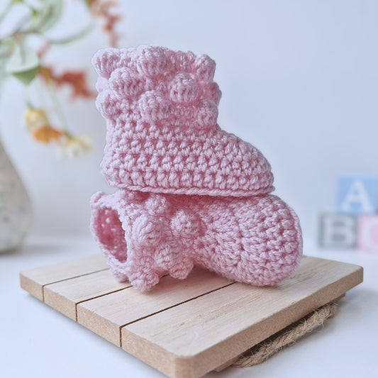 A pair of handmade powder pink crochet baby booties with a decorative bobble-stitch cuff, displayed on a wooden stand.