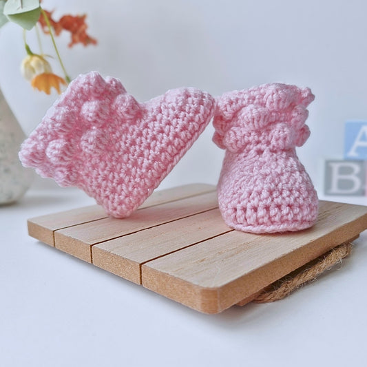 A pair of handmade powder pink crochet baby booties with a decorative bobble-stitch cuff, displayed on a wooden stand.