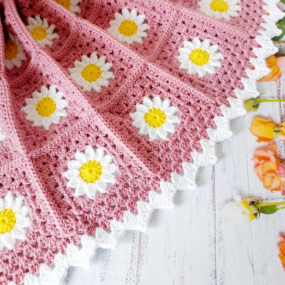 Pink crocheted blanket with white daisy patterns on a wooden surface