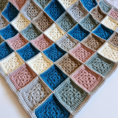 A handmade crocheted baby blanket featuring a patchwork of colorful square blocks, including shades of blue, pink, beige, brown, and gray, laid flat on a white surface.