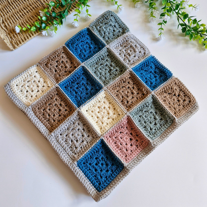 A handmade crocheted square blanket made of multiple textured granny-square panels in muted blue, brown, beige, pink, and gray tones, laid flat on a white surface with decorative greenery in the corners.