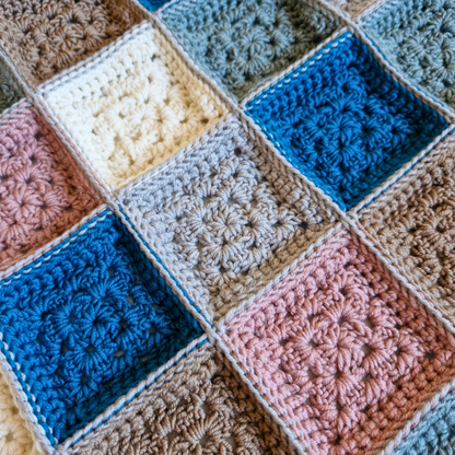 A close-up of a crocheted patchwork blanket with alternating textured squares in blue, pink, cream, taupe, and light gray.