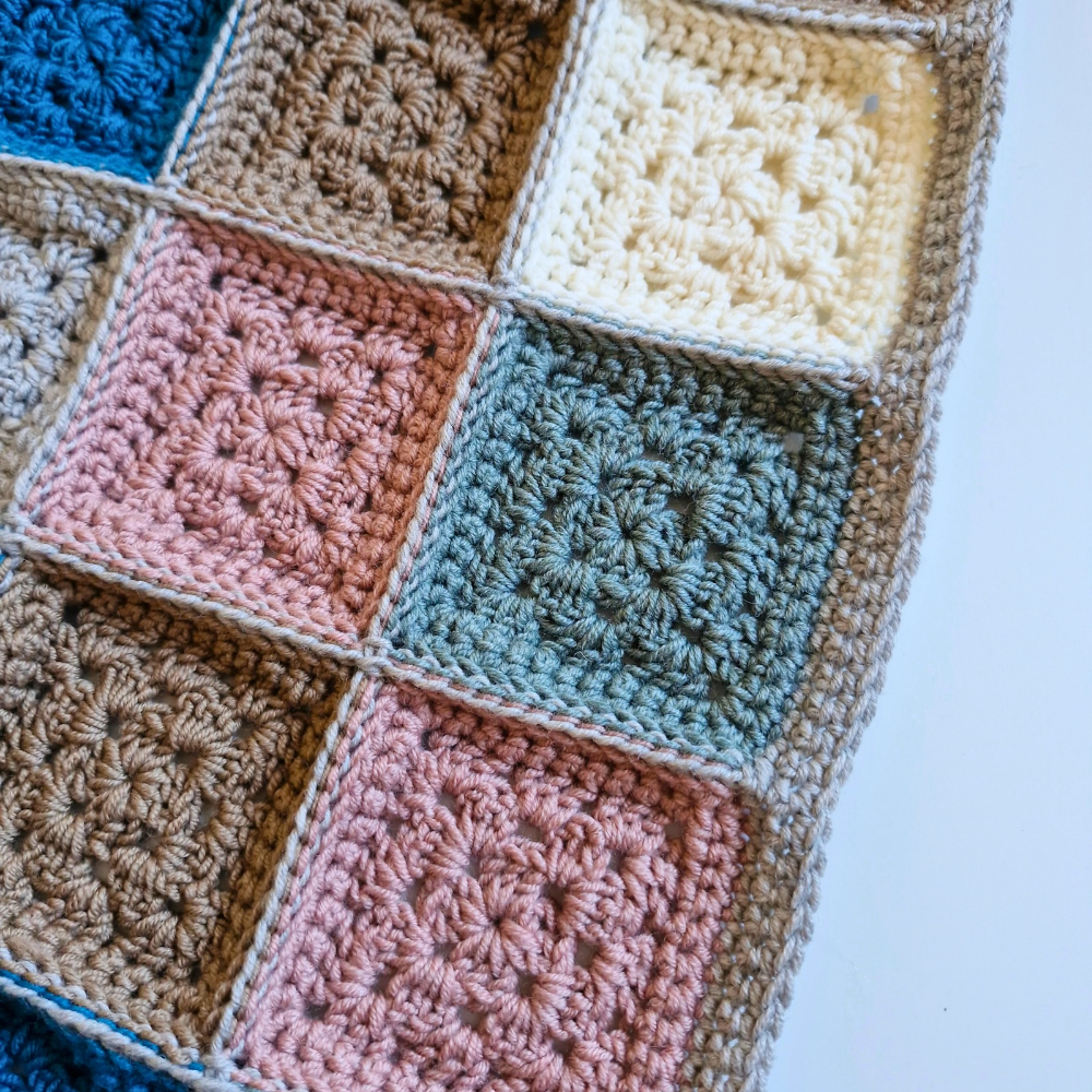 A close-up of a handmade crochet blanket featuring multiple square patches in muted colors including dusty pink, sage green, cream, tan, and blue, bordered by a thick beige crochet edge.