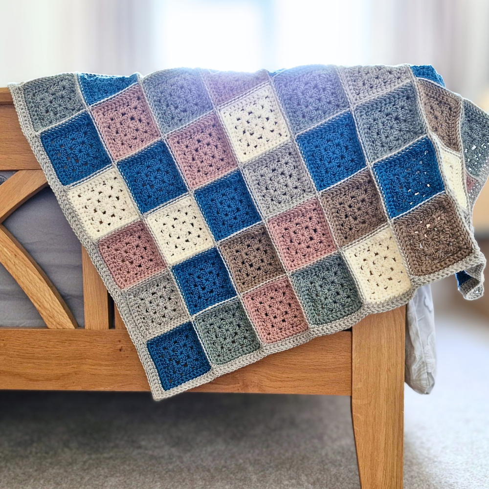A handmade crocheted blanket with a patchwork of colorful granny squares draped over a wooden chair in a softly lit room.