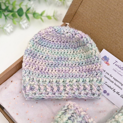 A pastel crocheted beanie hat displayed inside an open kraft gift box with a branded card, alongside a matching small accessory. The hat has soft variegated stripes in lavender, mint, pale blue, cream, and light purple.