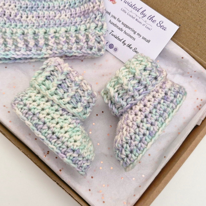 A handmade crochet baby bootie set in pastel variegated yarn, presented in a cardboard gift box with a handwritten thank you card. The set includes two booties and a matching hat.