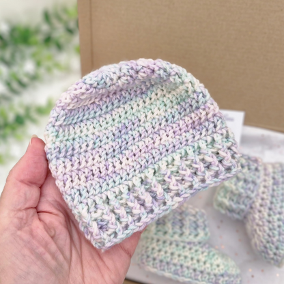 A hand holds a small crocheted beanie in soft pastel stripes of lavender, mint, cream, and pale blue. More matching beanie pieces are visible in the background, with a cardboard box and blurred greenery behind them.