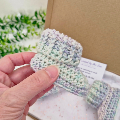 A hand holds a small crocheted baby bootie in pastel variegated yarn, with another matching bootie and a kraft gift box in the background.