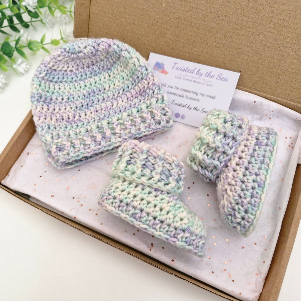 A handmade crochet baby bootie and hat set in a kraft gift box with a branded card. The crochet items are made in a variegated pastel yarn with shades of mint green, lavender, and cream.