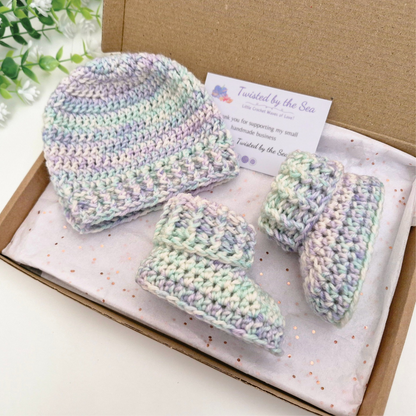 A handmade crochet baby bootie and hat set in a kraft gift box with a branded card. The crochet items are made in a variegated pastel yarn with shades of mint green, lavender, and cream.