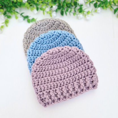 Three crocheted newborn baby hats in grey, blue, and pink on a white background with green leaves.