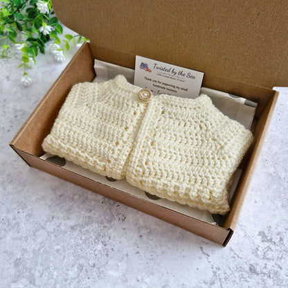 A cream-coloured crocheted baby cardigan with a single wooden button, presented inside a brown cardboard gift box with a branded card, set on a light gray stone surface.