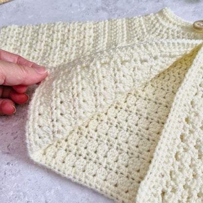A close-up of a cream-colored crocheted cardigan or sweater being held open by a hand, showing its textured knit pattern and button closure.