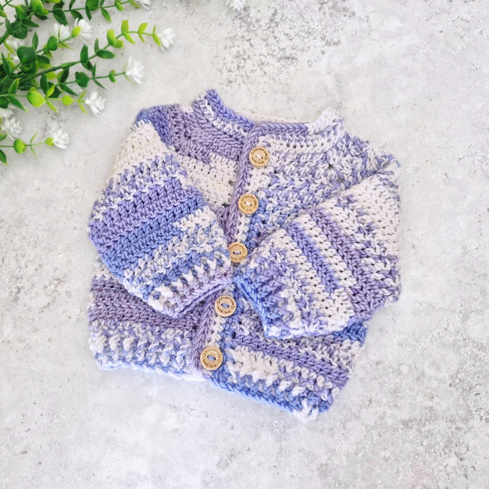 A handmade crochet baby cardigan sweater in lavender, light purple, and cream stripes, styled flat on a light gray textured surface with green and white flowers in the top-left corner.