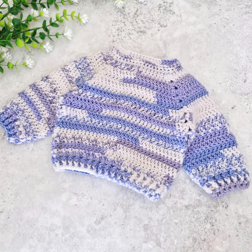 The backview of A handmade crocheted baby sweater with horizontal stripes in lavender, light blue, and cream, styled on a textured gray surface with green leaves in the corner.