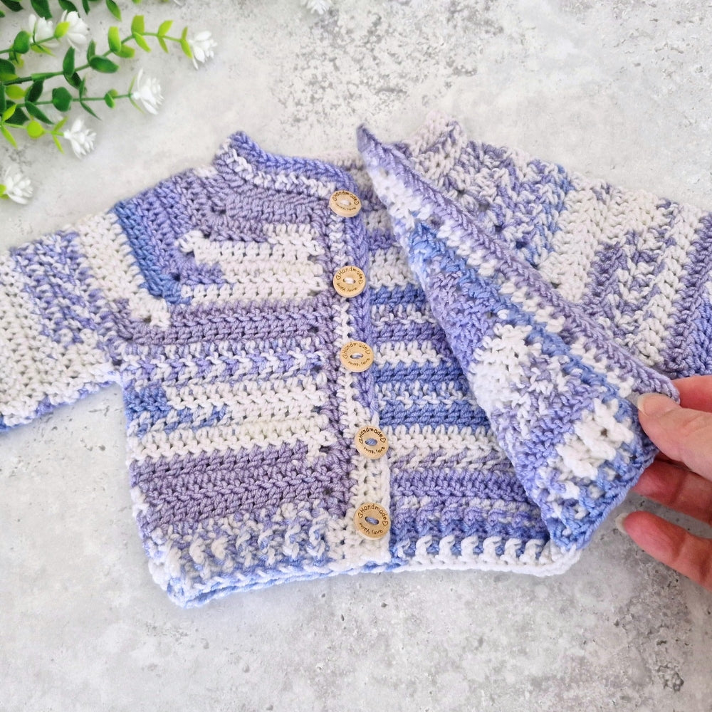 A handmade crocheted cardigan sweater for a baby, featuring lavender and off-white stripes with wooden buttons, shown on a light gray textured surface with a hand holding one side.