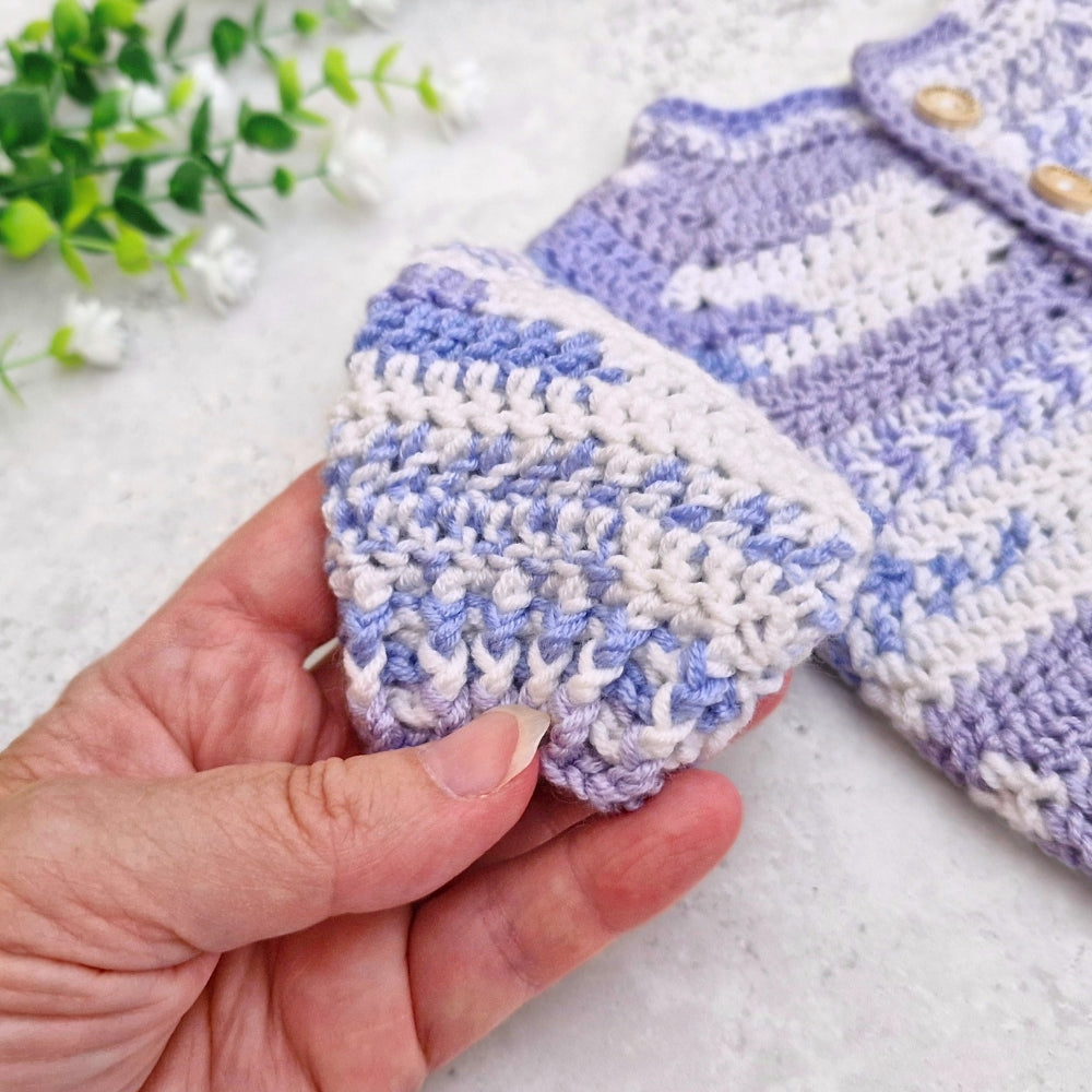 A close-up of a hand holding a crocheted baby cardifan sleeve in light purple and white yarn, with a matching crocheted garment laid on a white textured surface in the background.