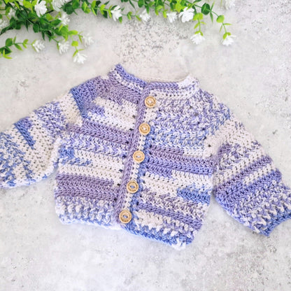 A handmade crochet cardigan sweater with lavender and off-white stripes, featuring wooden buttons down the front, laid flat on a textured light gray surface with decorative white flowers in the top corner.