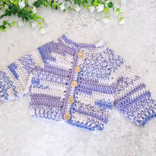A handmade crochet cardigan sweater with lavender and off-white stripes, featuring wooden buttons down the front, laid flat on a textured light gray surface with decorative white flowers in the top corner.