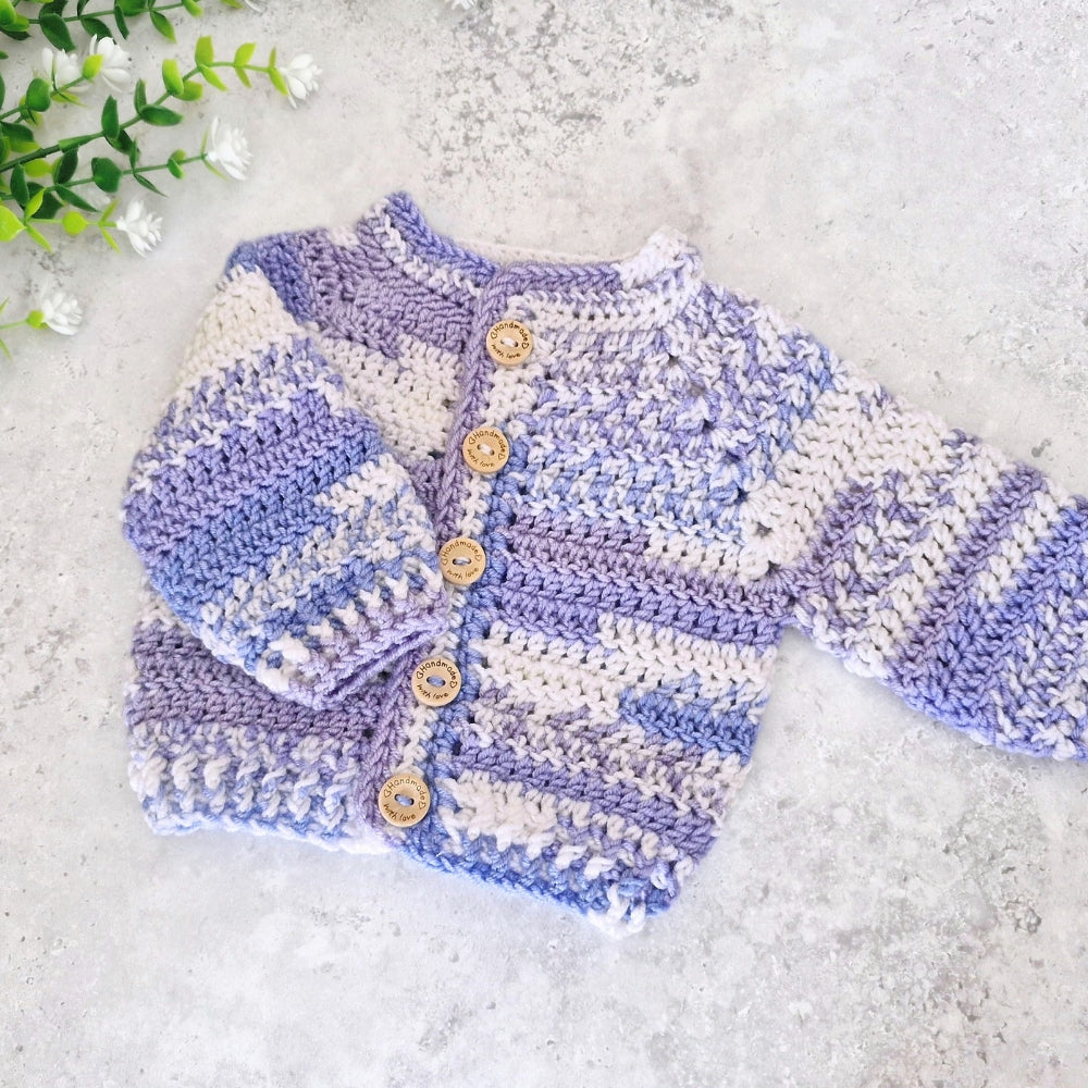 A handmade crocheted cardigan sweater for babies, featuring horizontal stripes of lavender purple, light blue, and cream. It has five wooden buttons down the front and is laid flat on a light gray textured surface with small white flowers in the top-left corner.