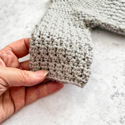 A close-up of a hand holding a light gray crocheted sleeve or cuff against a light stone-like background.