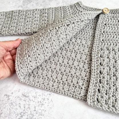 A close-up of a light gray crocheted cardigan or sweater with a textured bobble stitch pattern, shown with a hand holding the sleeve to demonstrate flexibility. The cardigan features a gold button closure and is laid flat on a light gray stone-textured surface.