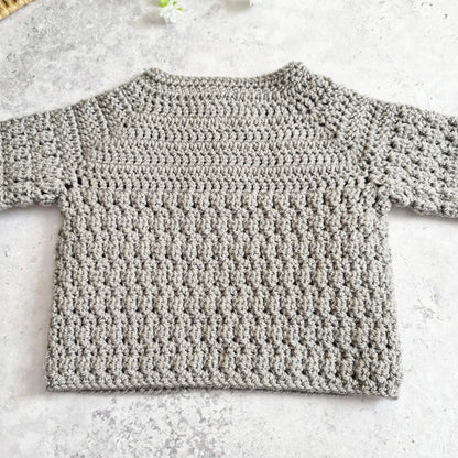 A light gray handmade crochet knit sweater with long sleeves, laid flat on a textured light gray surface.