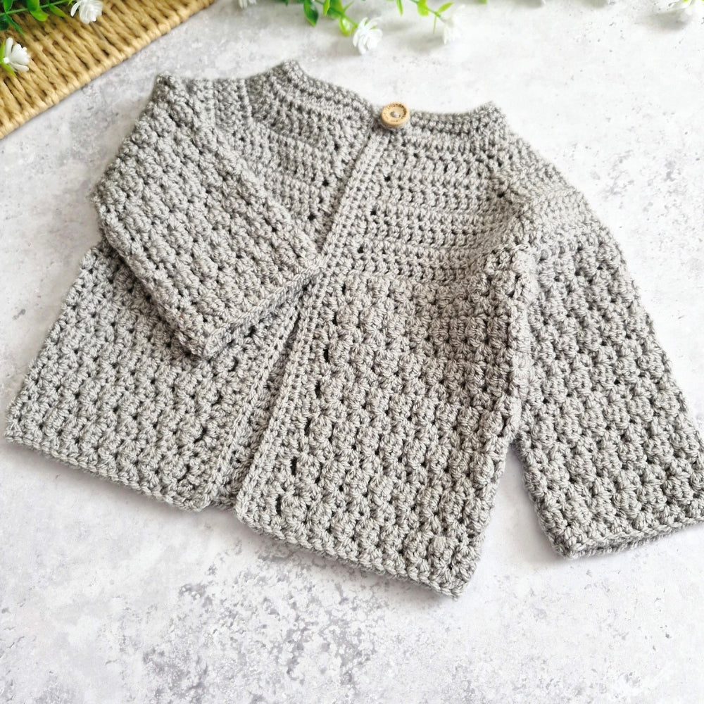 A light grey handmade crochet cardigan with long sleeves and a wooden button closure, laid flat on a textured light gray surface.