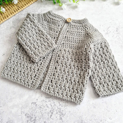 A light grey handmade crochet cardigan with long sleeves and a wooden button closure, laid flat on a textured light gray surface.