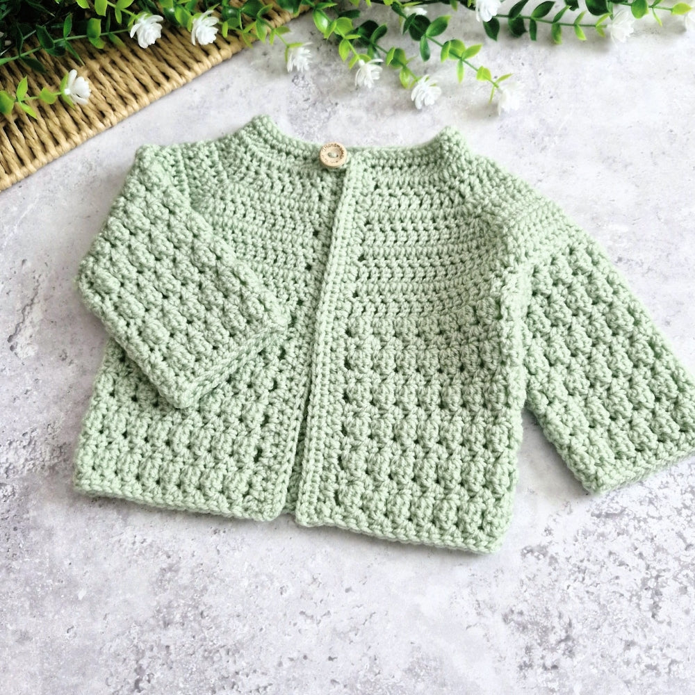 A light tea green crocheted cardigan sweater for a baby, featuring a textured open-knit pattern, long sleeves, and a single wooden button closure, laid flat on a light gray stone surface with decorative green leaves and a woven mat in the corner.