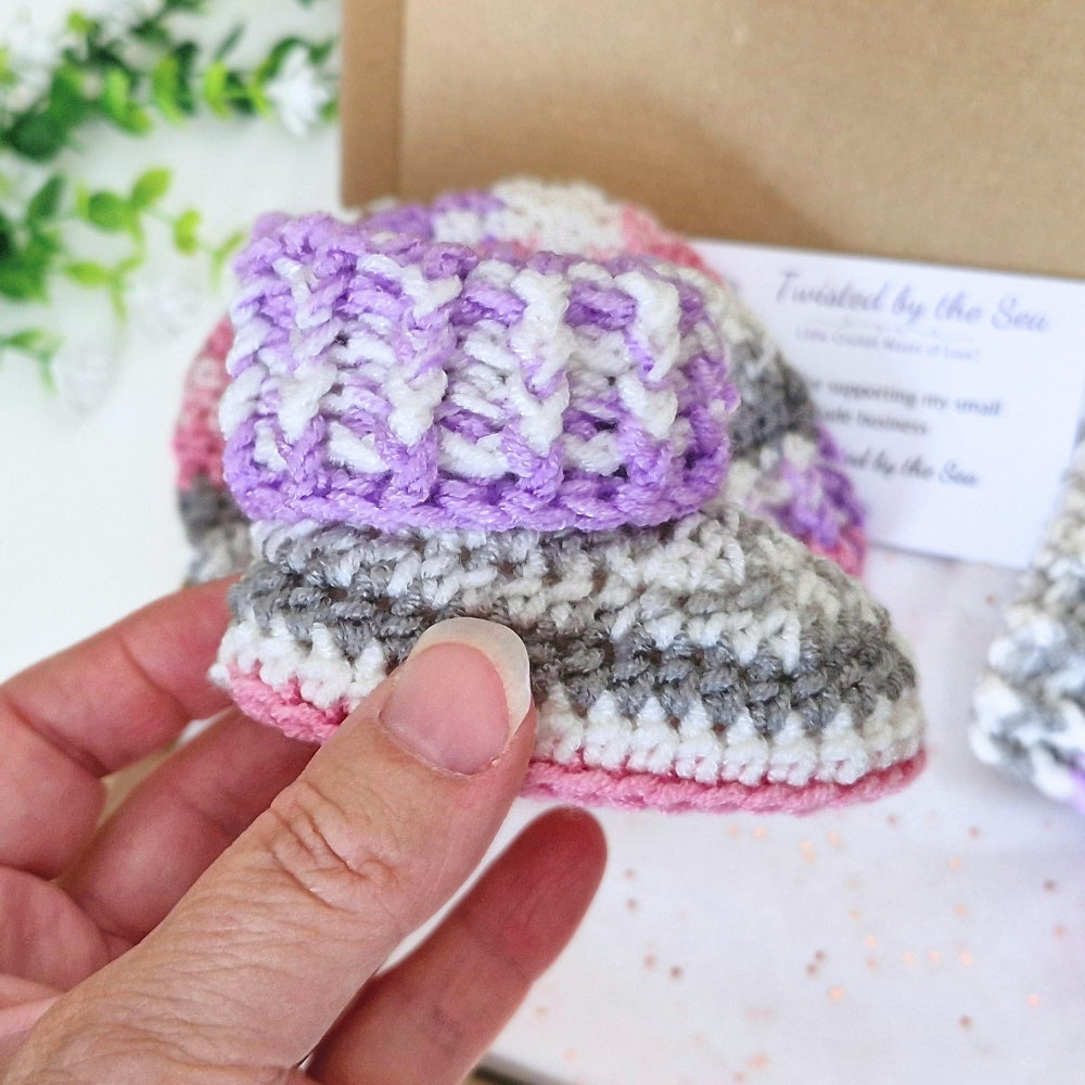 A hand holds a newborn handmade crocheted bootie in pastel colors, featuring a lavender and white knit cuff, gray and white striped body, and a pink edge, with a kraft box and care card in the background.