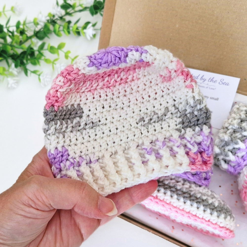 A hand holds a crocheted knit hat in pastel variegated colors of cream, pink, purple, and gray, with more matching booties and a kraft box in the background.